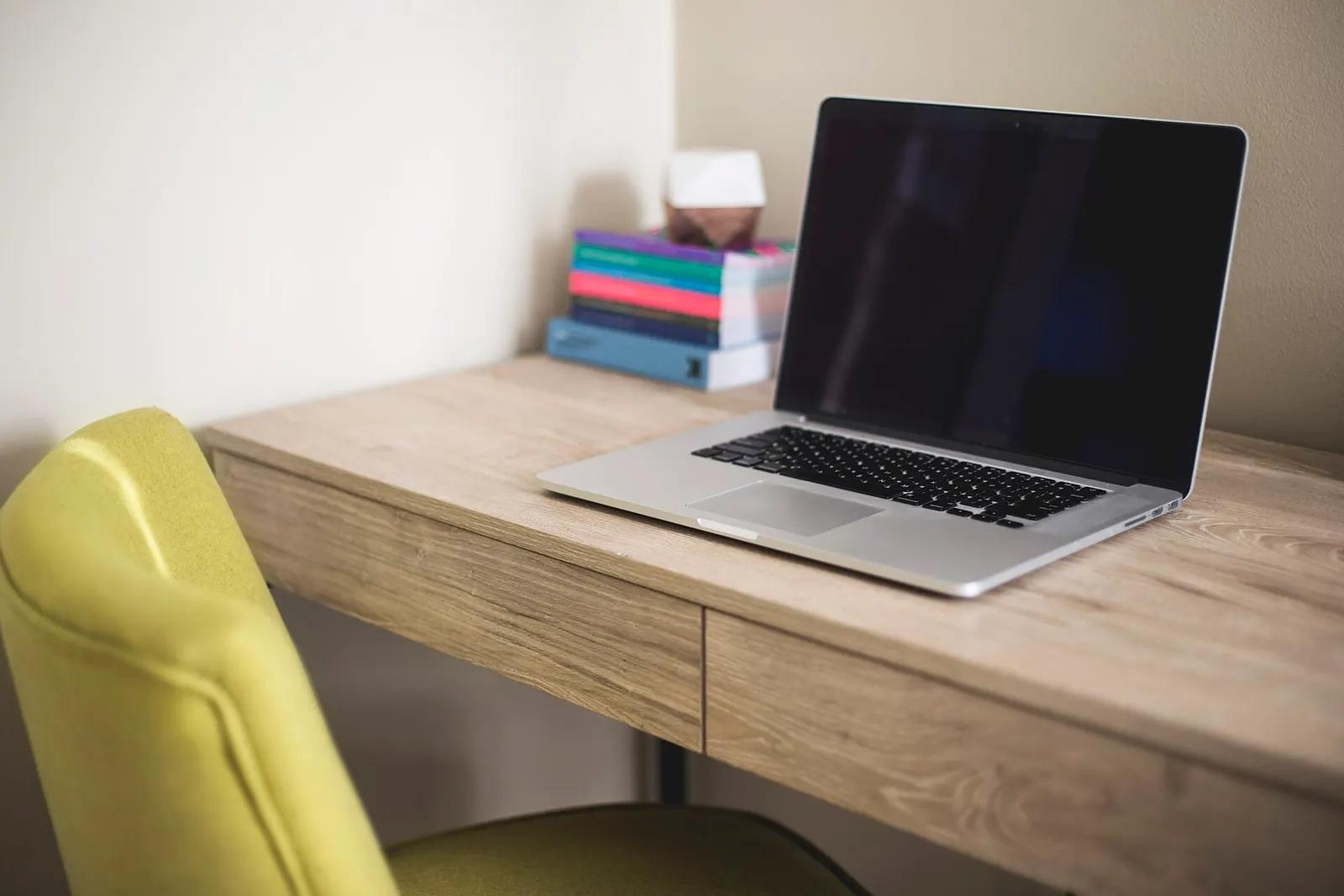 An open laptop on a small wooden desk beside a stack of books and a yellow chair
