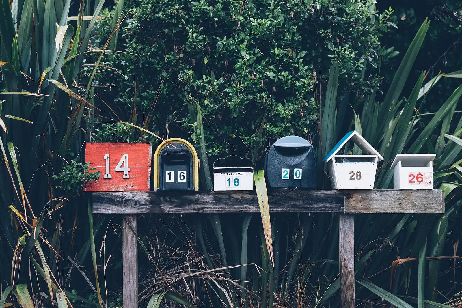 A row of six numbered mailboxes on a wooden rail in front of dense green plants