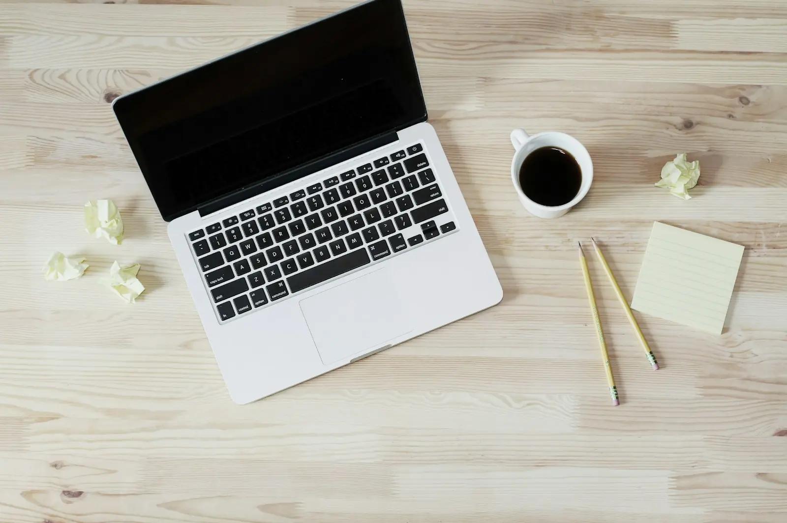 A laptop, a coffee cup and a notepad on a wooden desk seen from above