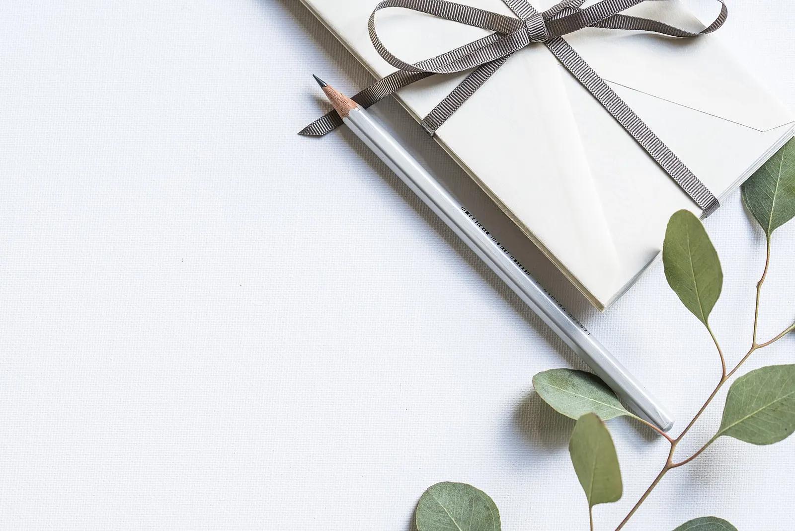 A bundle of cream envelopes tied with a grey ribbon next to a pencil and eucalyptus leaves